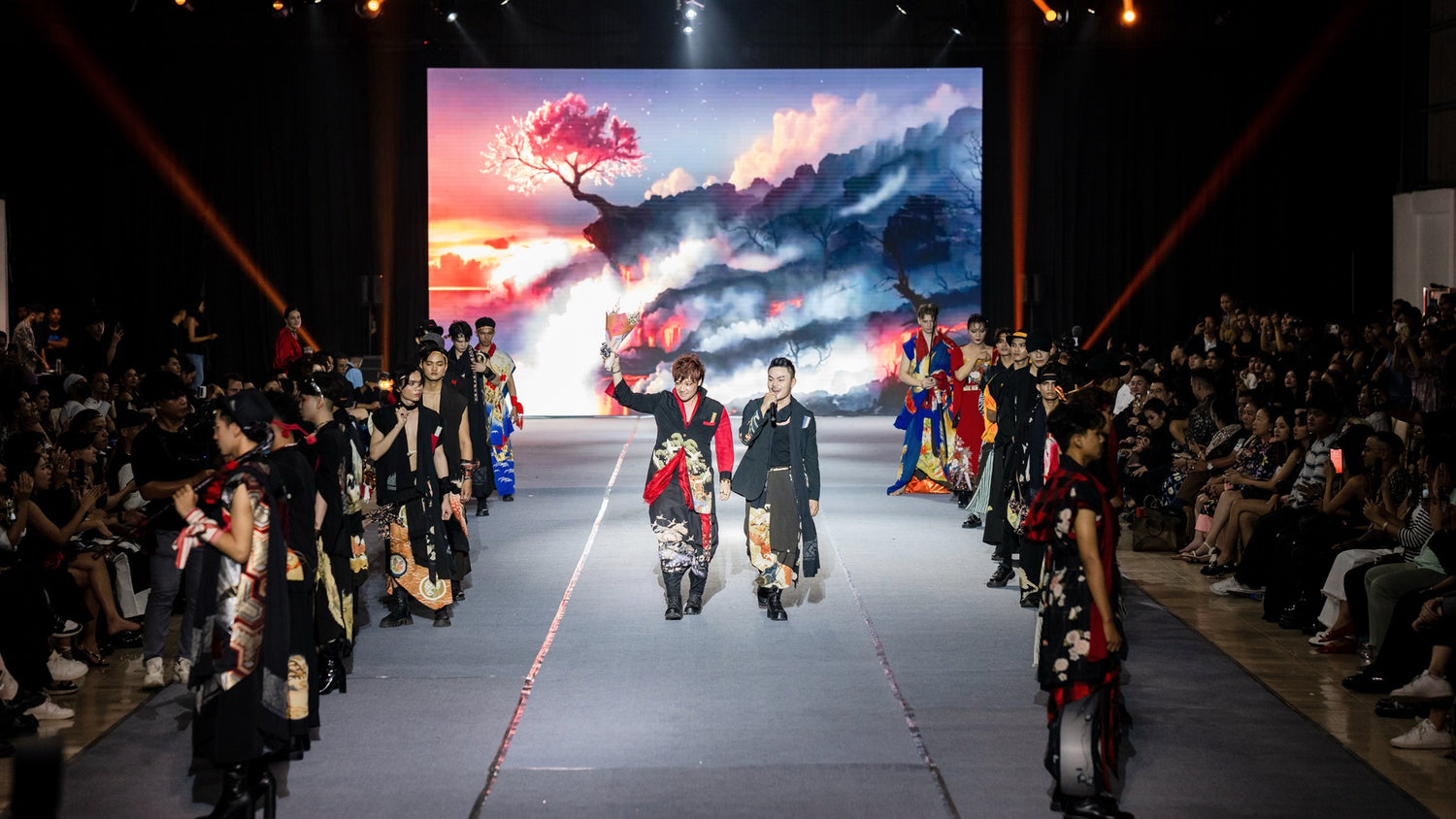 TATZU designer on stage holding a bouquet at the finale of last year’s fashion show, surrounded by the full lineup of models.